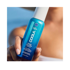Bilde av COOLA Classic Face Mist SPF 50 i elegant flaske, i en hånd på stranden, som fremhever produktets solbeskyttende og hudpleiende egenskaper